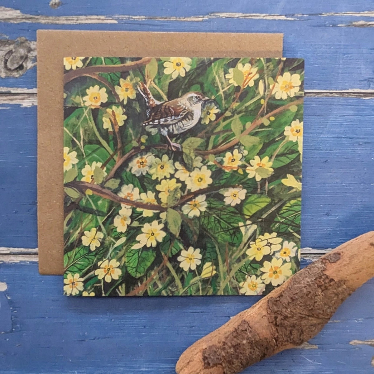 A square art card featuring a wren perched among green leaves and small yellow primrose flowers, placed in front of a brown kraft envelope on a distressed blue wooden surface with a piece of driftwood in the corner.
