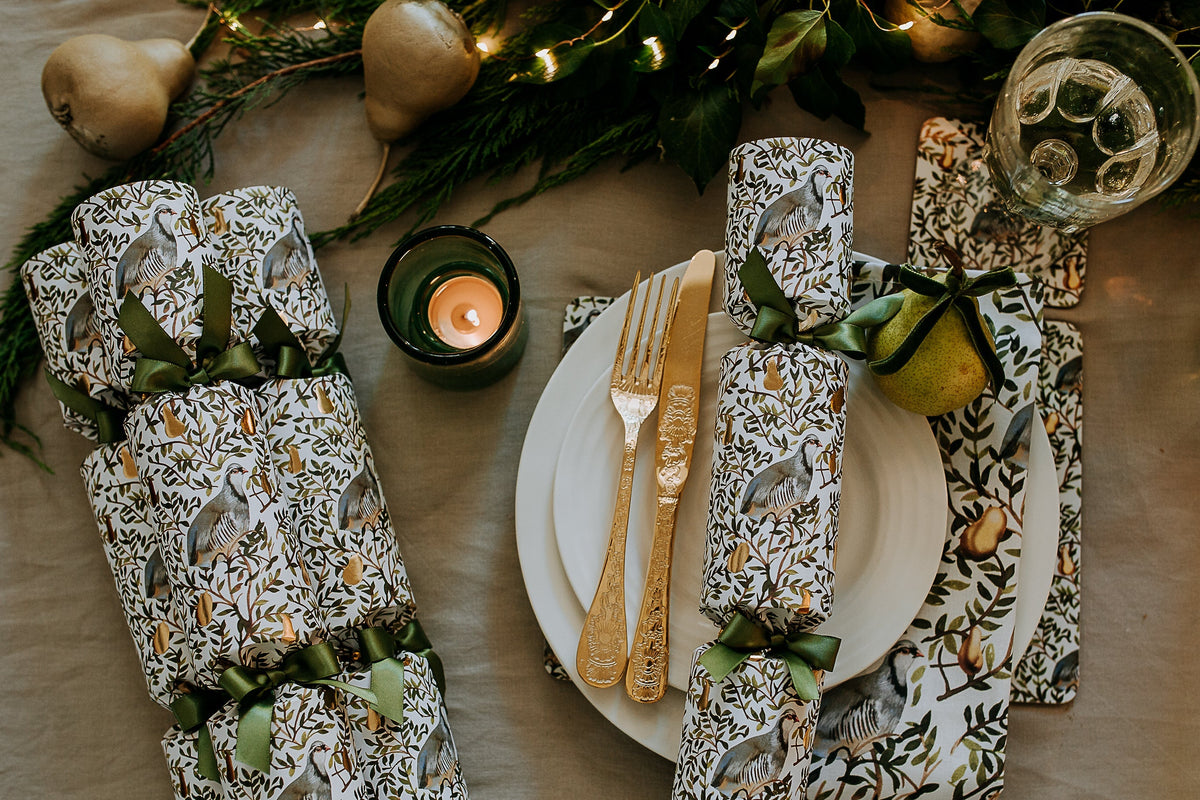 A styled holiday table setting featuring  Christmas crackers with a partridge in a pear tree pattern and green ribbons, alongside a white plate with gold-toned cutlery, a lit candle, and festive greenery with pear ornaments.