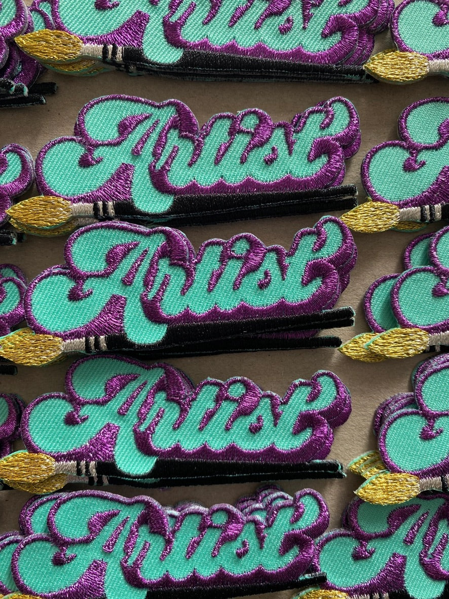 A close-up of multiple embroidered iron on patches  featuring the word 'Artist' in a stylised teal script with a purple glittery outline, each decorated with a small gold paintbrush at the bottom, arranged on a neutral surface.
