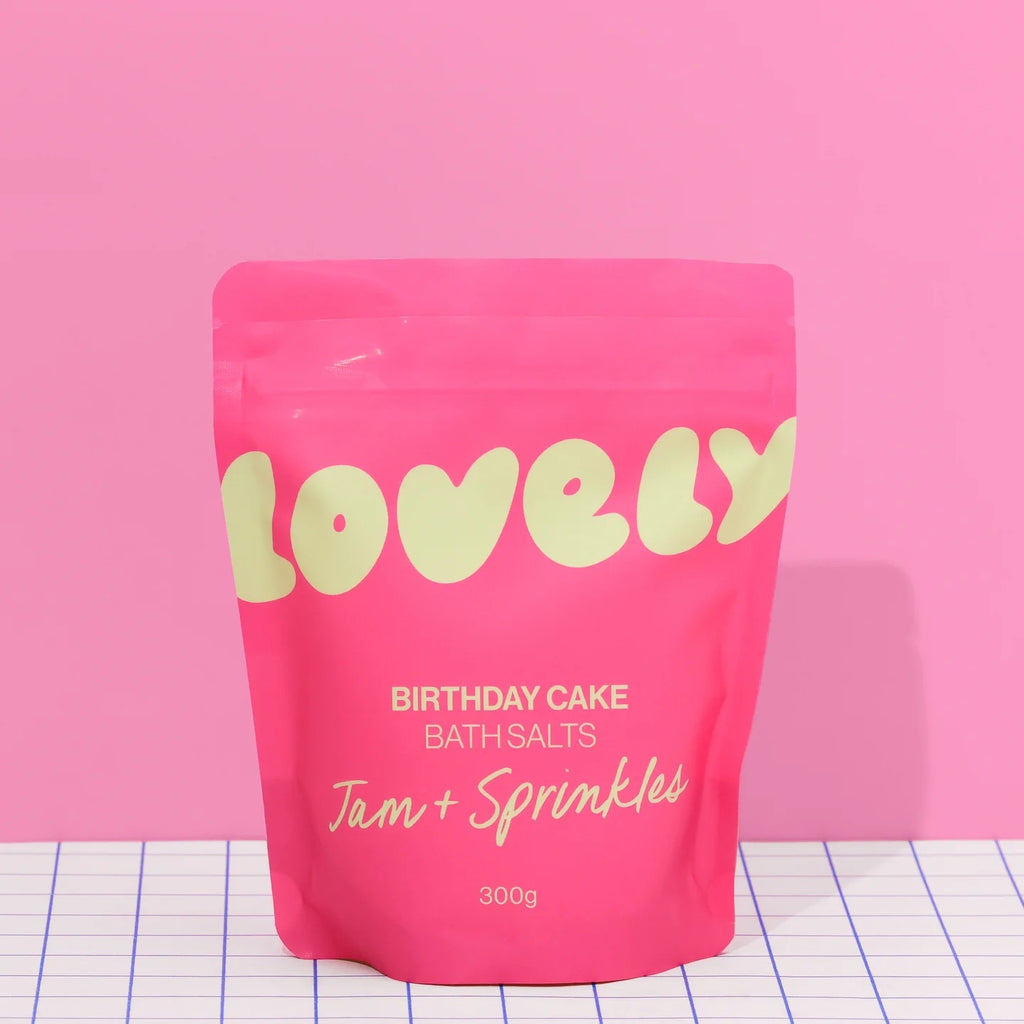 A bright pink pouch of Lovely Skincare's Birthday Cake Bath Salts, The pouch is on a white grid-patterned surface against a solid pink background.