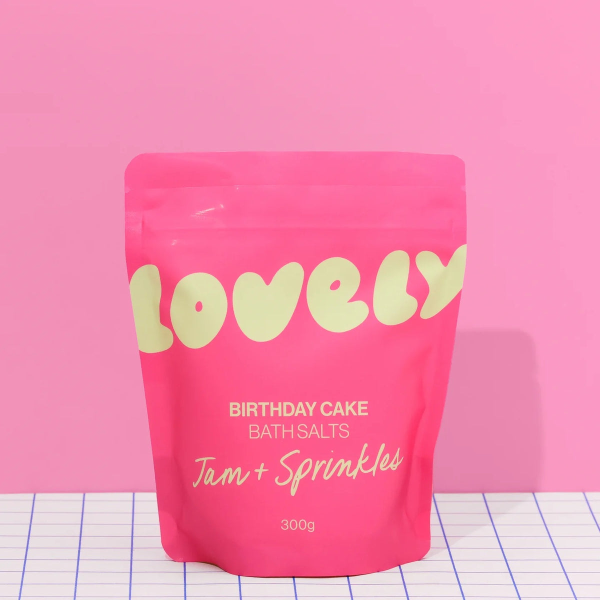 A bright pink pouch of Lovely Skincare's Birthday Cake Bath Salts, The pouch is on a white grid-patterned surface against a solid pink background.