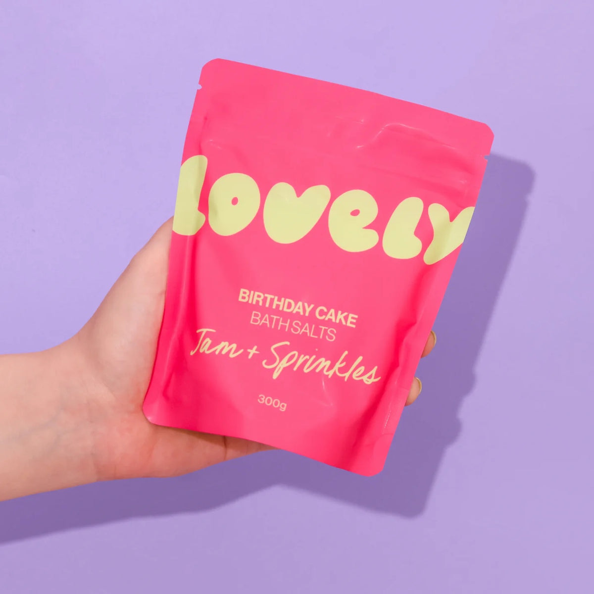 Hand holding a pink packet of 'Birthday Cake' bath salts on a purple background