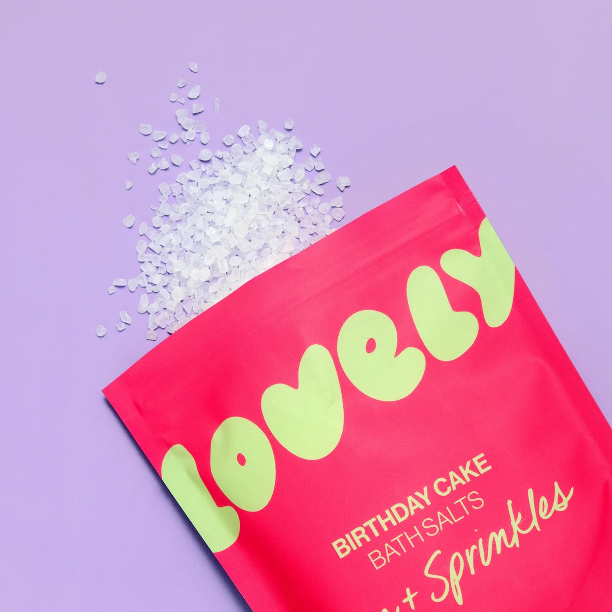 A pouch of bath salts, shown on a lavender background with white bath salts spilled from the top. The text reads 'BIRTHDAY CAKE BATH SALTS' and 'Jam + Sprinkles'.