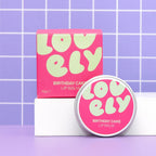 A pink tin of Lovely Skincare's Birthday Cake Lip Balm with its matching pink box, displayed on a white block against a purple tiled background.