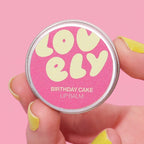 Lip balm by Lovely Skincare, Birthday Cake flavour held by a hand with yellow nail polish against a pink background.