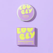 Lovely Skincare's Blueberry Cheesecake Lip Balm shown with its round tin and matching square box on a solid lavender background. The packaging features a light purple background with bold, playful yellow text spelling 'LOVELY' and smaller yellow text indicating the flavor.