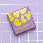 A square box of Lovely Skincare's Blueberry Cheesecake Lip Balm, featuring bold yellow lettering, placed on a light purple grid background.