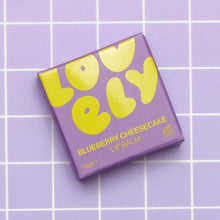 A square box of Lovely Skincare's Blueberry Cheesecake Lip Balm, featuring bold yellow lettering, placed on a light purple grid background.