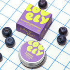 A box of Lovely Skincare's Blueberry Cheesecake Lip Balm arranged on a white surface with a blue grid pattern and scattered blueberries. The packaging is purple with bright yellow lettering.