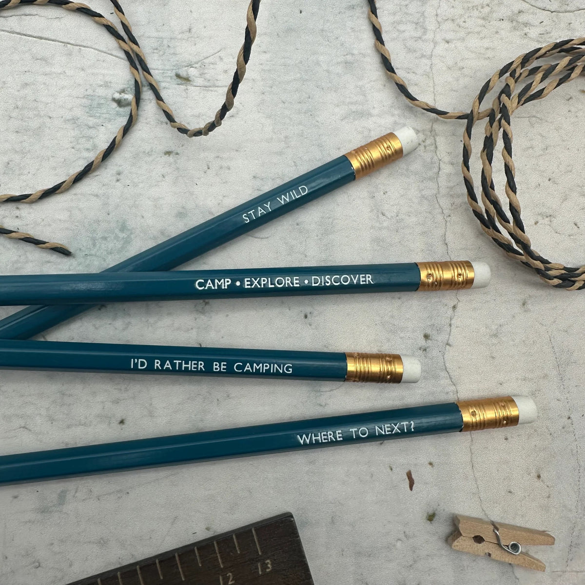 Four teal pencils with gold ferrules and white erasers, printed with camping-themed quotes, arranged on a light gray textured surface with burlap and a wooden clothes peg nearby.