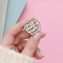 Hand holding an enamel pin with 'Can I go read my book?' text against a pink background