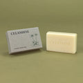 A cream-colored natural soap bar embossed with 'CELANDINE' sits next to its textured gray packaging box, both featuring the brand name 'CELANDINE' and a simple floral illustration, set against a solid olive green background.