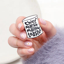 Hand holding an enamel pin in shape of marshmallow with text reading 'Chief Marshmallow Toaster' in black on a white background.