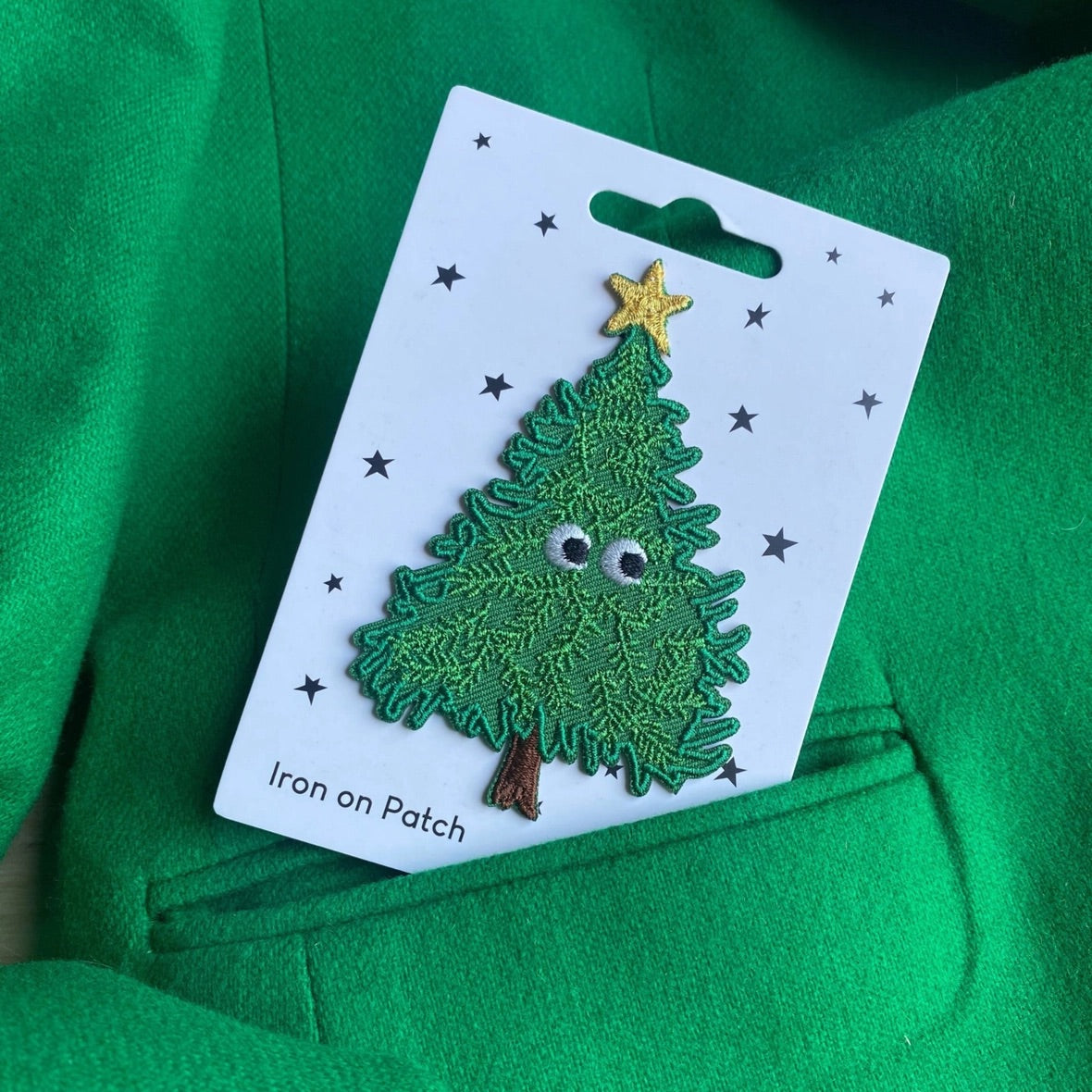 An embroidered Christmas tree patch on a white backing card, placed on a bright green fabric surface. The patch is green with a yellow star on top, small gray eyes, and a brown trunk.