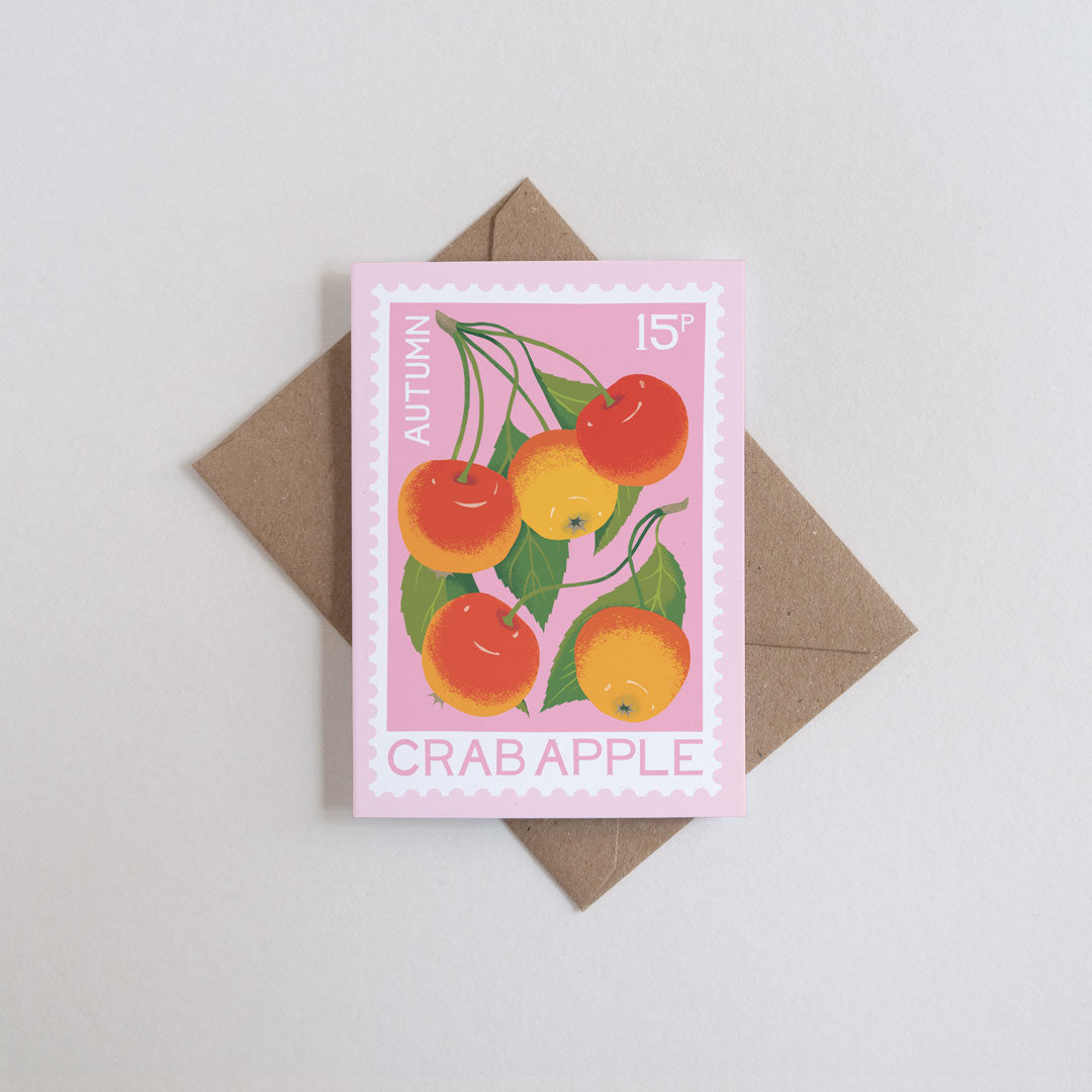 A pink stamp-style card featuring an illustration of crab apples in autumn colors, placed on top of a brown envelope against a light gray background.