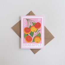 A pink stamp-style card featuring an illustration of crab apples in autumn colors, placed on top of a brown envelope against a light gray background.