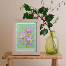 A framed art print featuring purple crocus flowers on a mint green background, styled on a wooden table next to a green glass vase with holly branches against a beige wall.