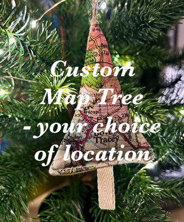 A fabric Christmas tree ornament made from vintage-style map print material hangs on a green Christmas tree branch with warm string lights. White text overlay reads 'Custom Map Tree - your choice of location'.