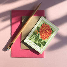 A flat-lay of a mini greeting card featuring a Dahlia flower, placed on top of a brown envelope and a bright pink card, with a wooden pencil resting diagonally on a pink surface under soft shadows.