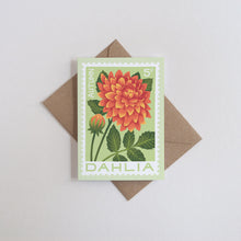 A rectangular greeting card featuring an illustrated orange and yellow dahlia flower with green leaves on a pale green background, placed on top of a brown envelope against a light gray surface.