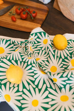 Tea towel close up with white and yellow daisies on a dark green background, styled with lemons and tomatoes.