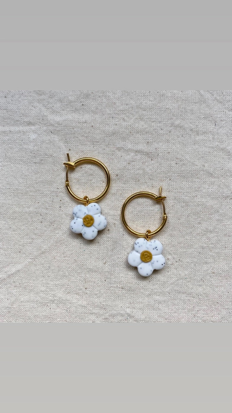 A pair of gold hoop earrings with dangling white daisy charms, displayed on a textured beige fabric background.