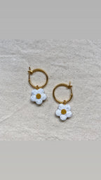 A pair of gold hoop earrings with dangling white daisy charms, displayed on a textured beige fabric background.