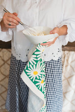 A person in a white blouse and blue-and-white gingham skirt holds a white bowl while whisking. A green kitchen towel with large white daisy flowers hangs from her hands showing yellow flower centers.