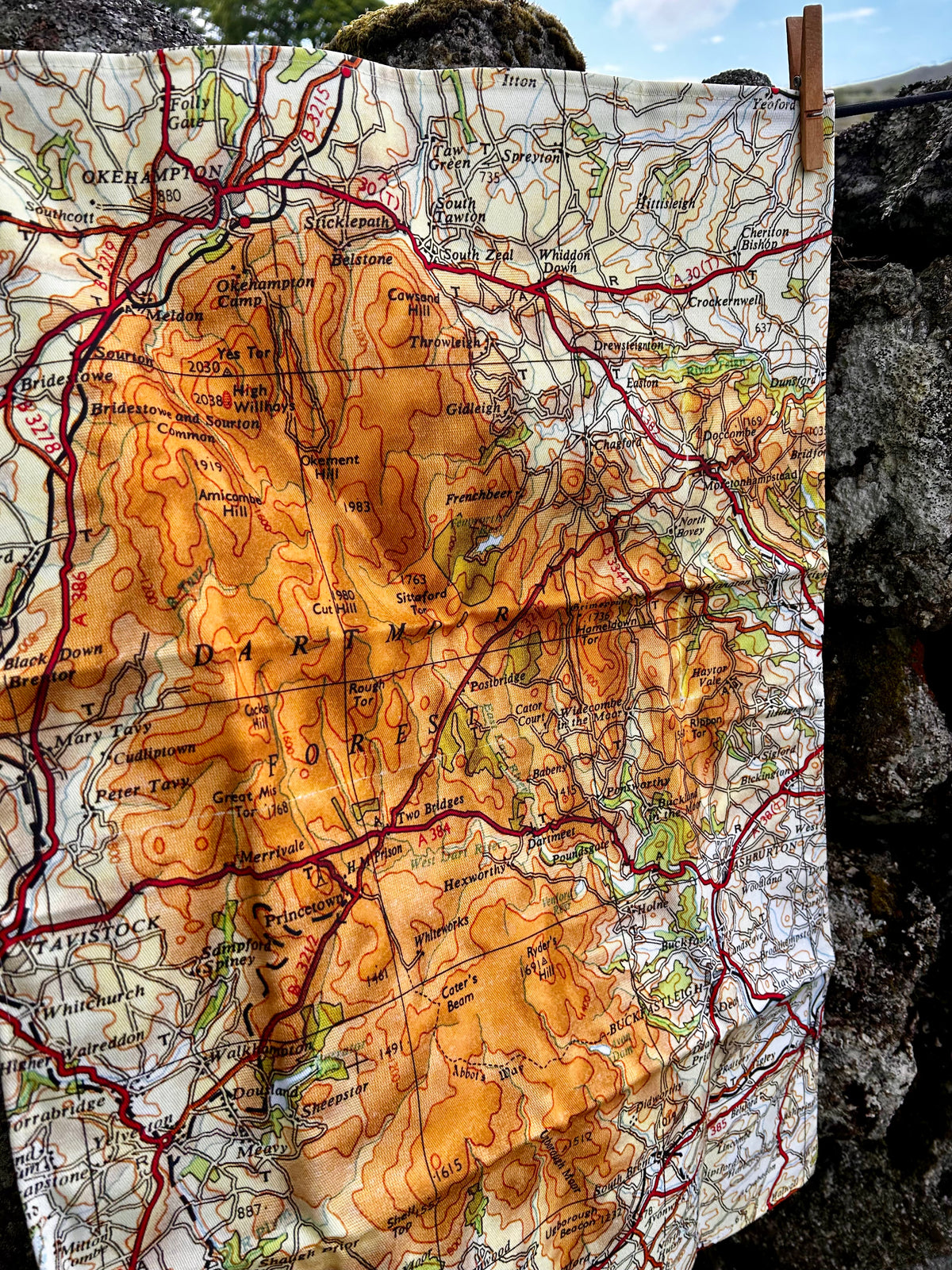 A map tea towel of Dartmoor, pegged against a stone wall outdoors. The map shows orange-brown shaded terrain, red road lines, and black text on an off-white background.