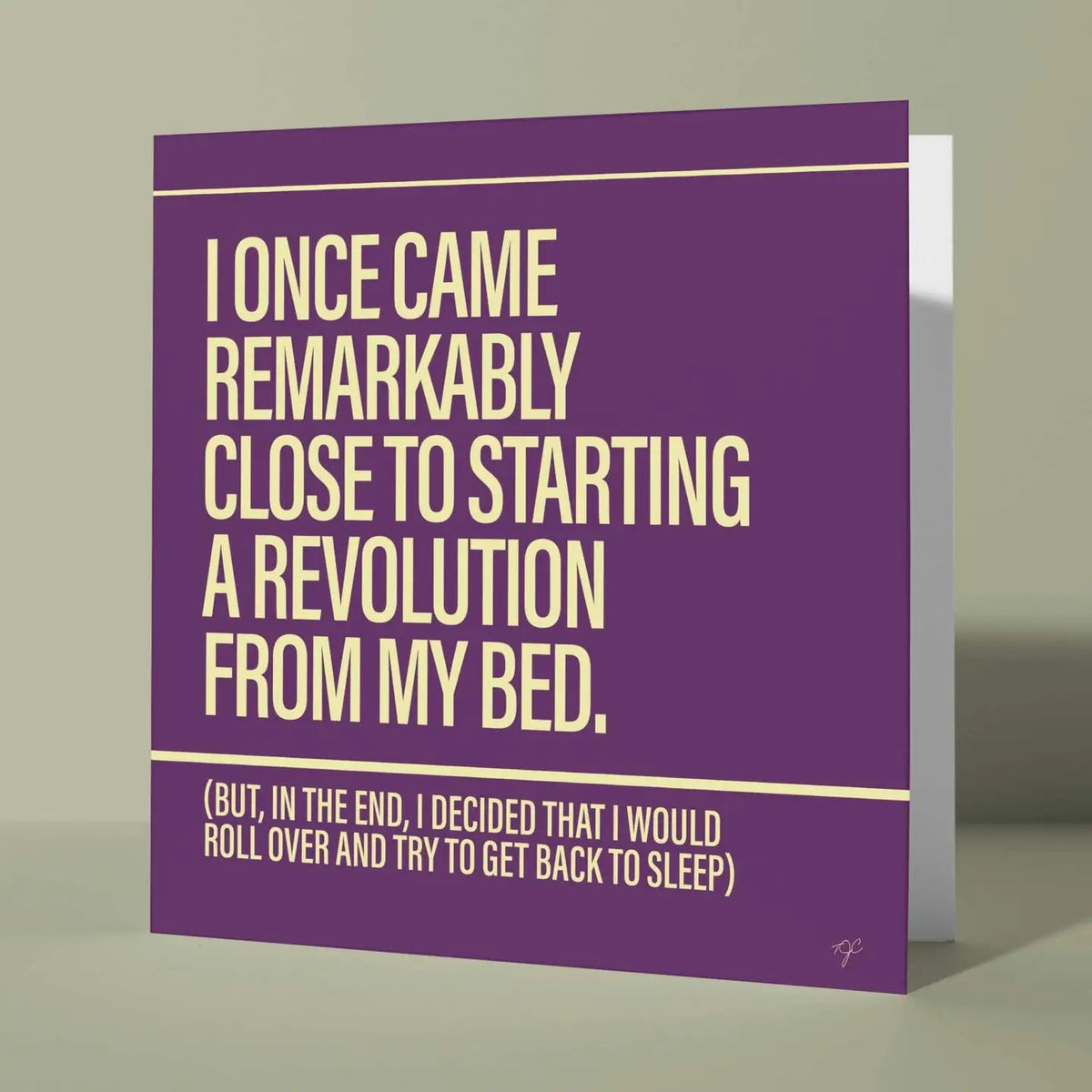 A purple greeting card with cream text that reads, 'I ONCE CAME REMARKABLY CLOSE TO STARTING A REVOLUTION FROM MY BED. (BUT, IN THE END, I DECIDED THAT I WOULD ROLL OVER AND TRY TO GET BACK TO SLEEP)' shown standing against a neutral beige background.