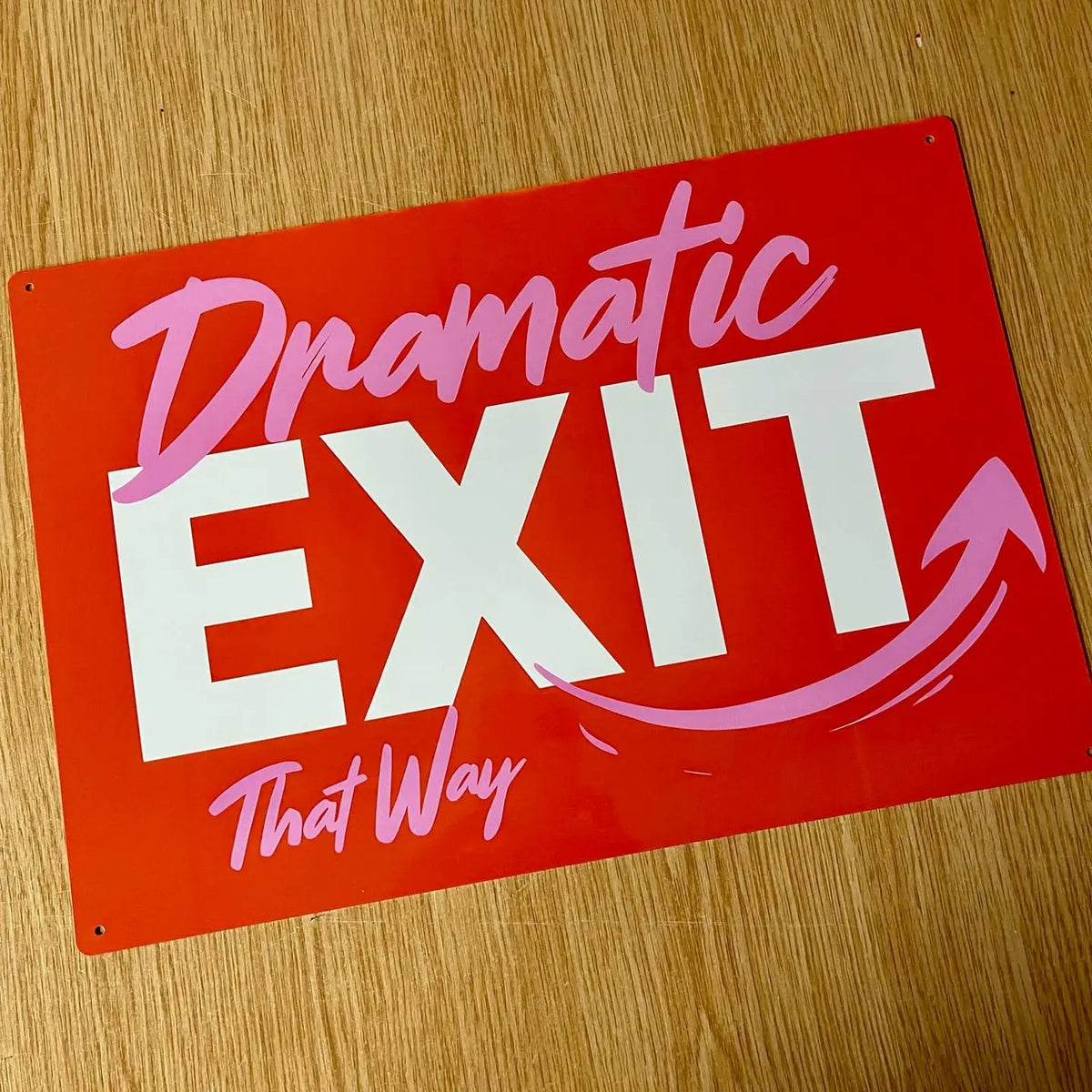 Red sign with 'Dramatic EXIT That Way' and arrow text in pink and white on a wooden surface