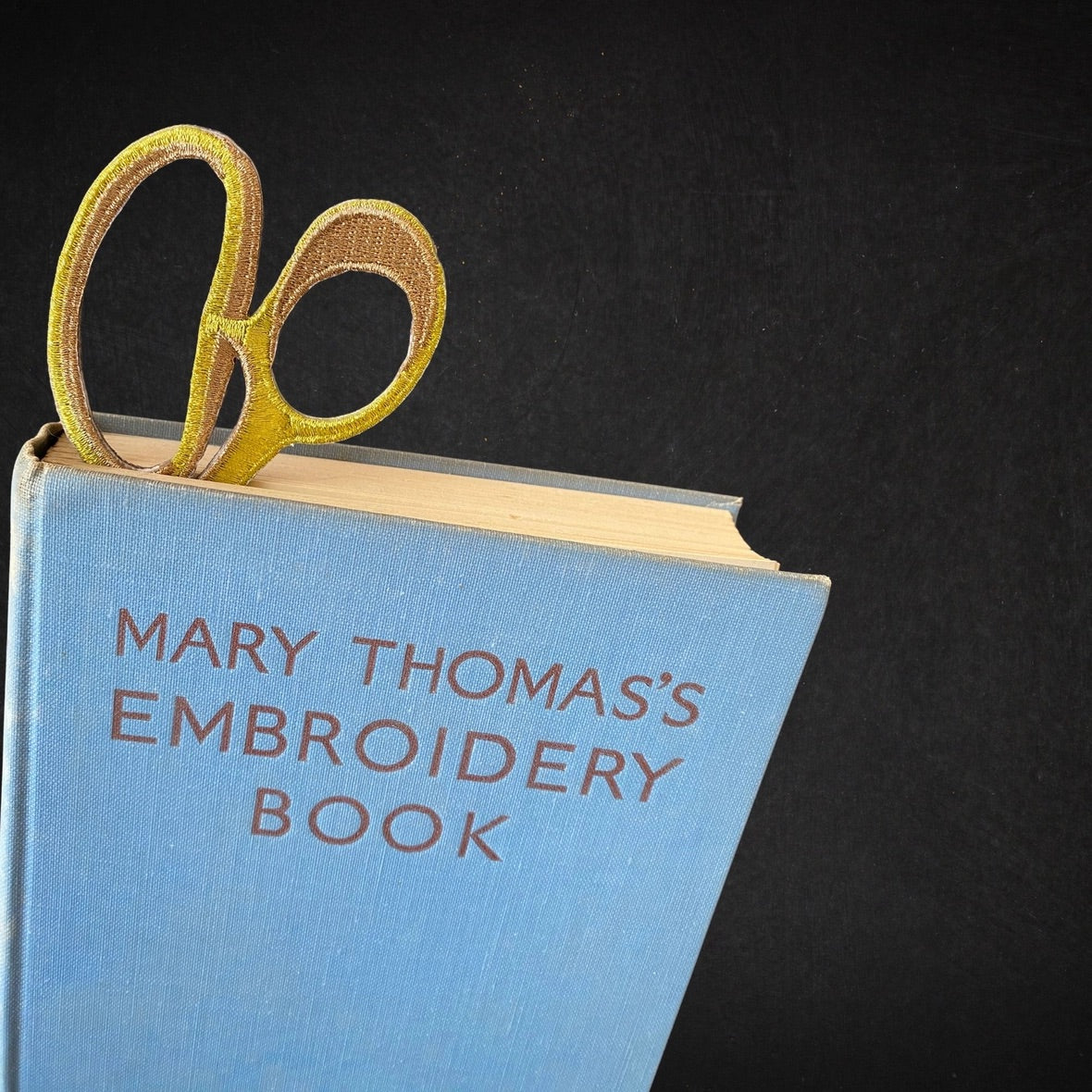 A light blue hardcover book titled 'MARY THOMAS'S EMBROIDERY BOOK' with gold embroidery, photographed on a black background with a gold pair of scissors embroidered bookmark tucked inside the book