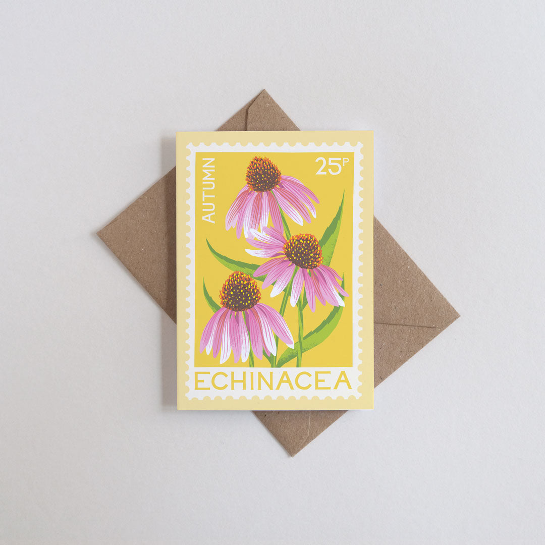 A rectangular greeting card featuring an illustration of three pink echinacea flowers with green stems on a yellow background. The card is placed on top of a brown envelope against a light gray background.