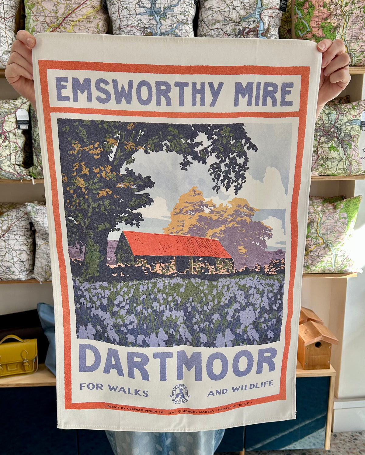 A person holds up a tea towel featuring a vintage-style 'Emsworthy Mire Dartmoor' landscape print with a red-roofed barn, trees, and purple wildflowers. 