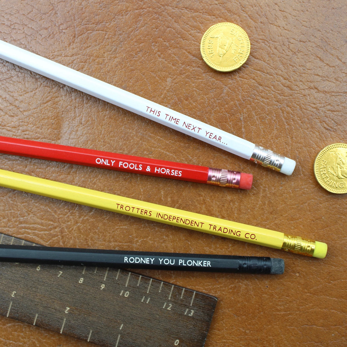 Four wooden pencils in white, red, yellow, and black are arranged on a brown textured leather surface alongside a wooden ruler and two gold coins. The pencils feature humorous quotes from the iconic sitcom 'Only Fools & Horses'.