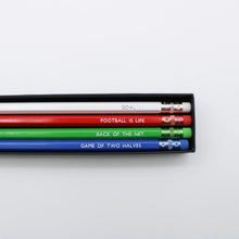 Four wooden pencils in white, red, green, and blue, displayed inside a black giftbox on a clean white background. Each pencil features a different football quote.