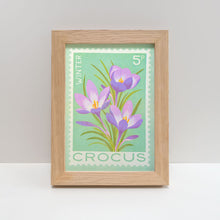 A framed art print featuring a vintage-style crocus stamp illustration with three purple flowers on a mint green background, set in a light natural wood frame.