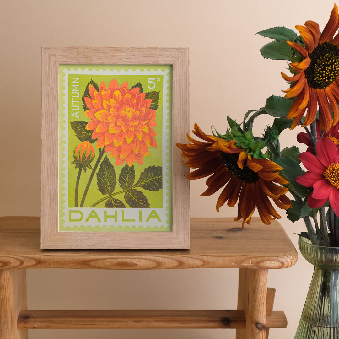 A framed art print featuring an orange and yellow dahlia flower on a light green background, styled on a wooden table next to a vase of sunflowers against a beige wall.
