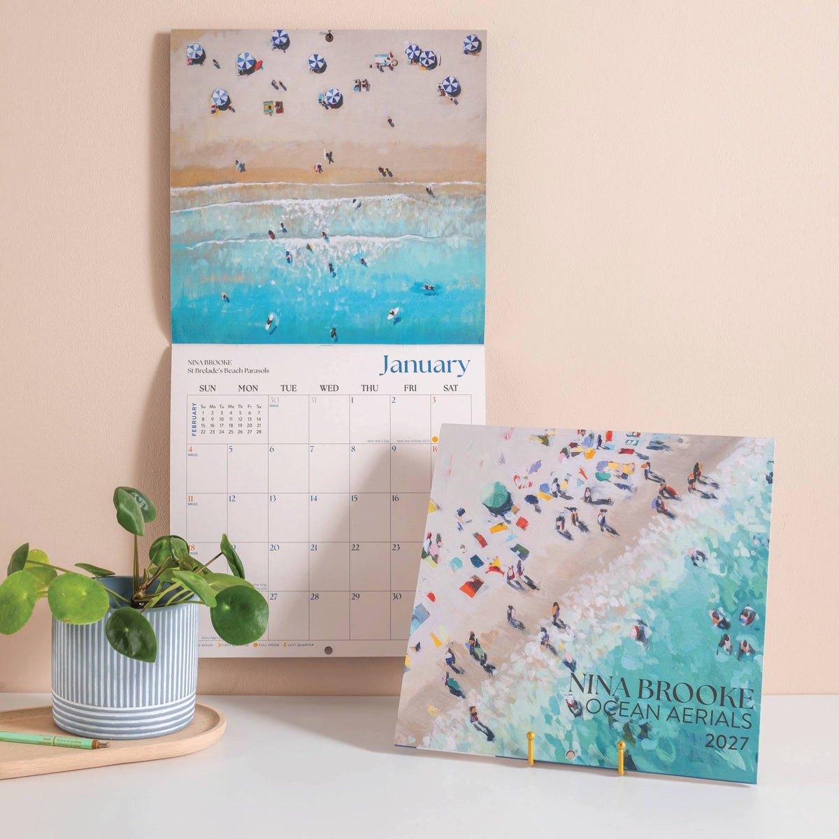 Open square calendar hung on wall , plus additional calendar shown standing featuring aerial paintings of the ocean and beach with pale peach wall and houseplant.