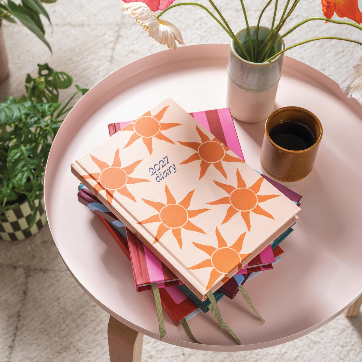 Stack of colourful diaries with sun designs on a pink table, surrounded by plants and a cup.
