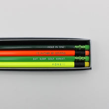 Four golf-themed pencils in a silver pencil case. The pencils are lime green, bright orange, dark green and neon yellow, each printed with a golf-related phrase.