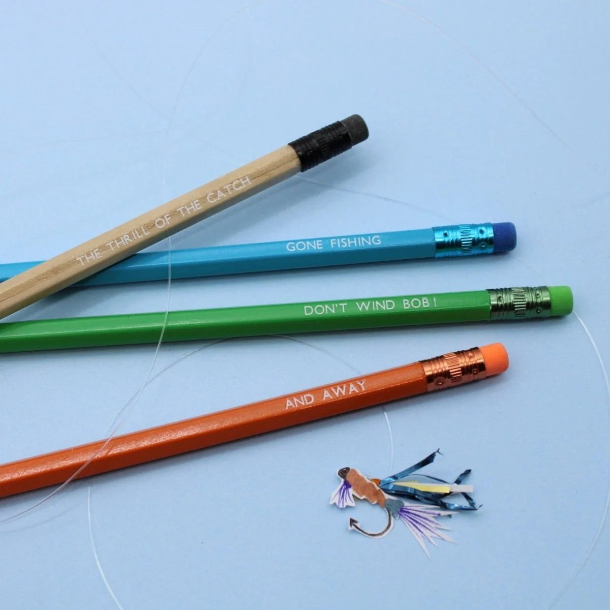 Four fishing-themed pencils in brown, blue, green, and orange are arranged on a light blue background, with a small fly fishing lure placed near the bottom right. The pencils feature Gone Fishing-related phrases engraved on them.