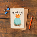 Greeting card with chicken and egg illustration and text reading 'You're such a good egg' on a wooden surface with a pen and paper clips.