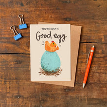 Greeting card with chicken and egg illustration and text reading 'You're such a good egg' on a wooden surface with a pen and paper clips.
