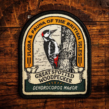An embroidered patch featuring a Great Spotted Woodpecker on a tree trunk, with the text 'FLORA & FAUNA OF THE BRITISH ISLES' and 'GREAT SPOTTED WOODPECKER' and 'DENDROCOPOS MAJOR'. The patch is displayed on a rustic wooden background.