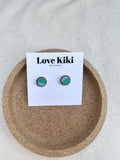 Circular green and blue polymer clay stud earrings with gold metal edge shown on white 'Love Kiki' branded backing card in cork dish