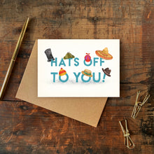 Greeting card with colorful hats and 'Hats Off To You!' text on a wooden surface with a pen and paper clips.