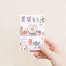 Enamel pin in pale copper colour on a white background with the text 'Hot Chocolate Connoisseur' and an illustration of a hot chocolate in the middle. Shown on a patterned backing card