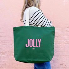 Person holding a green tote bag with 'JOLLY TIRED' printed on it against a pink background