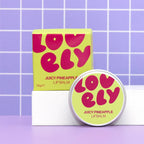 Lovely Skincare's Juicy Pineapple Lip Balm shown with its box and round tin container, set against a purple tiled background.